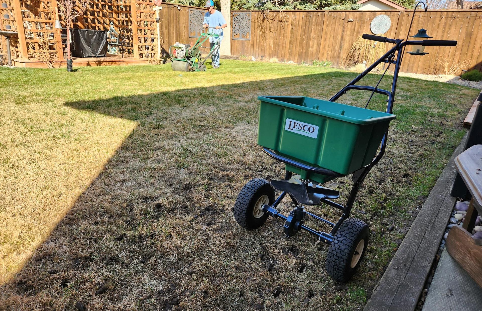 A green broadcast spreader sits on a patchy lawn with a person using a lawn aerator in the background.