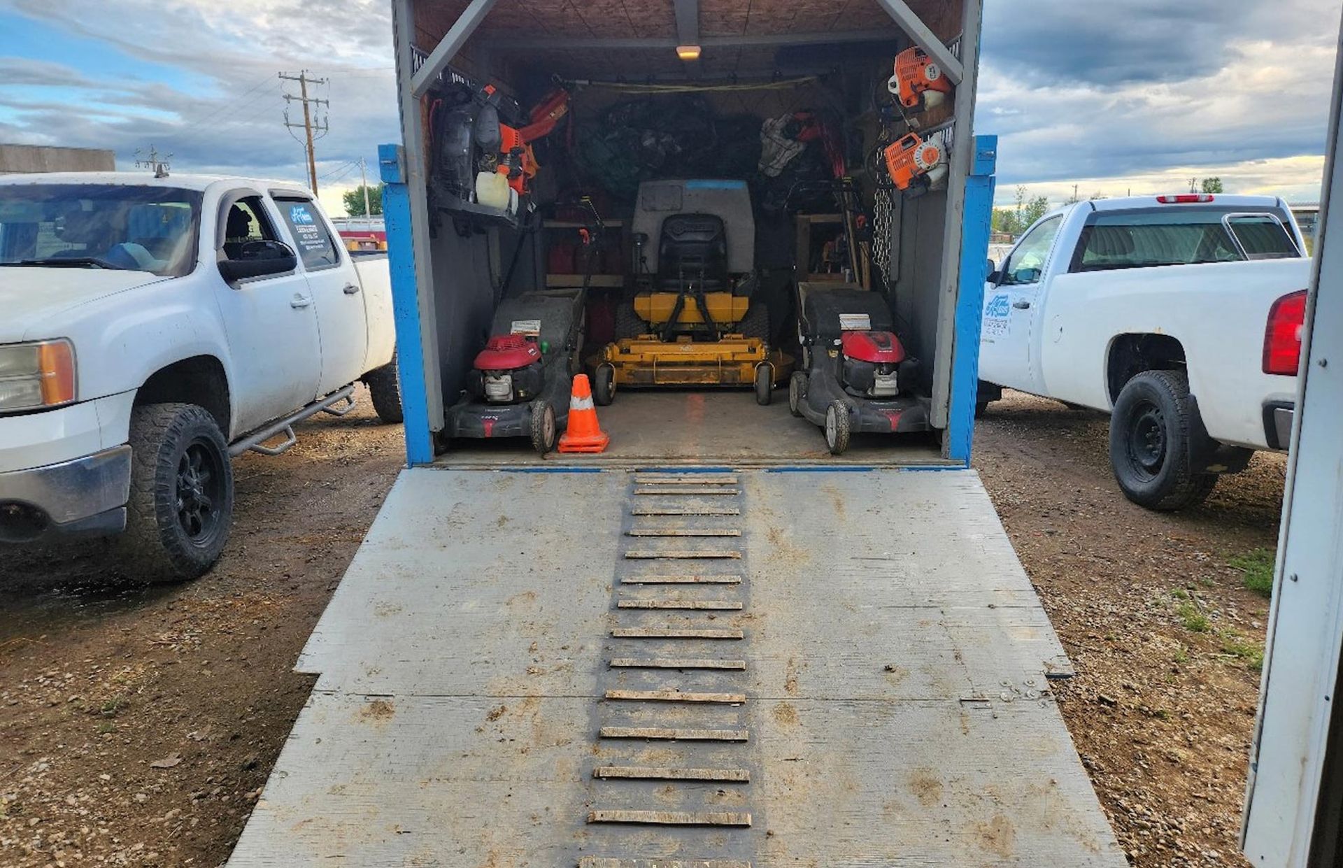 Rear view of a trailer containing two push mowers and a riding lawn mower, parked between two white pickup trucks.