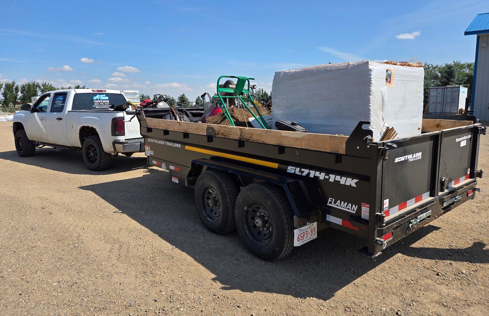 A white pickup truck towing a black dump trailer loaded with construction materials on a gravel lot under a blue sky.