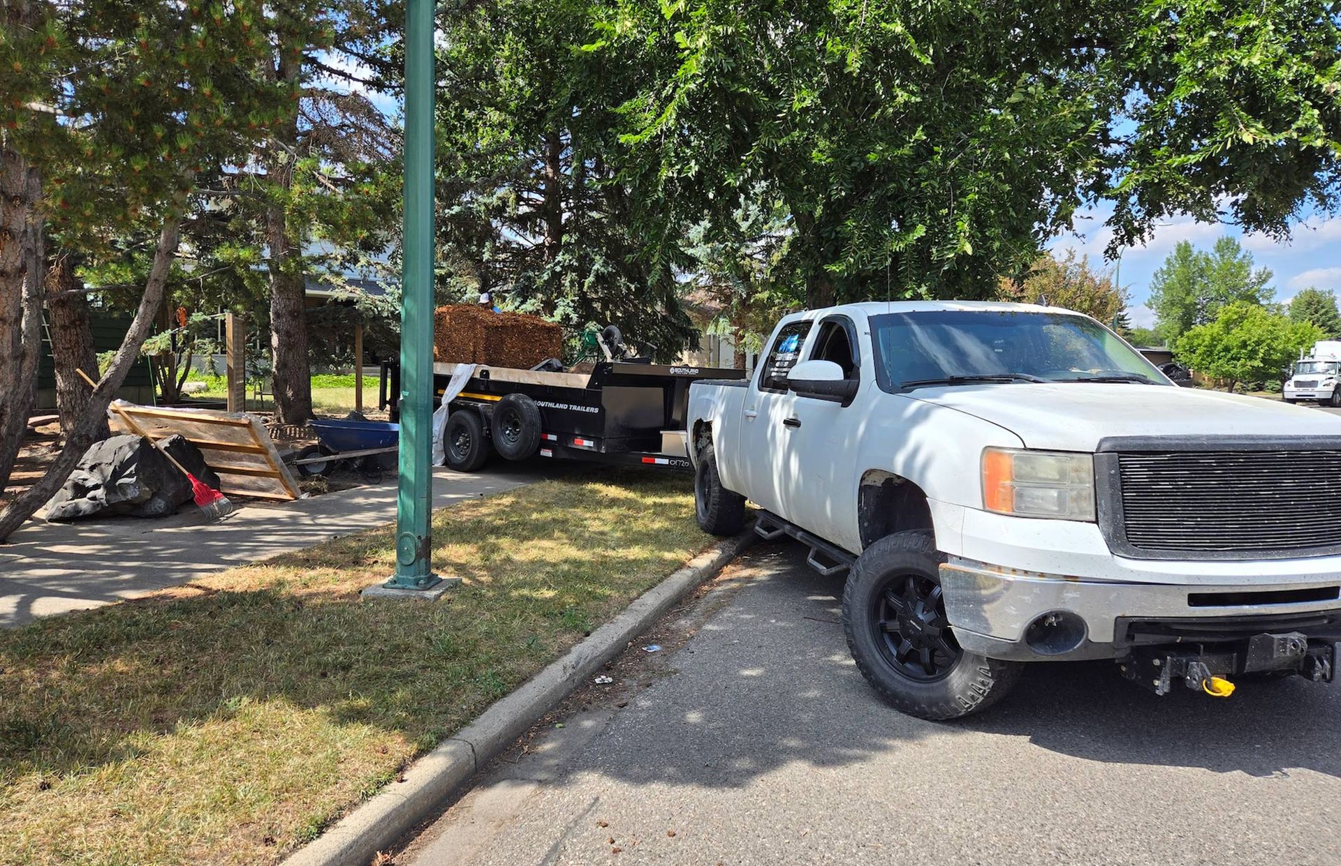 A white pickup truck with a trailer attached is parked on the side of a grassy residential street near some trees.