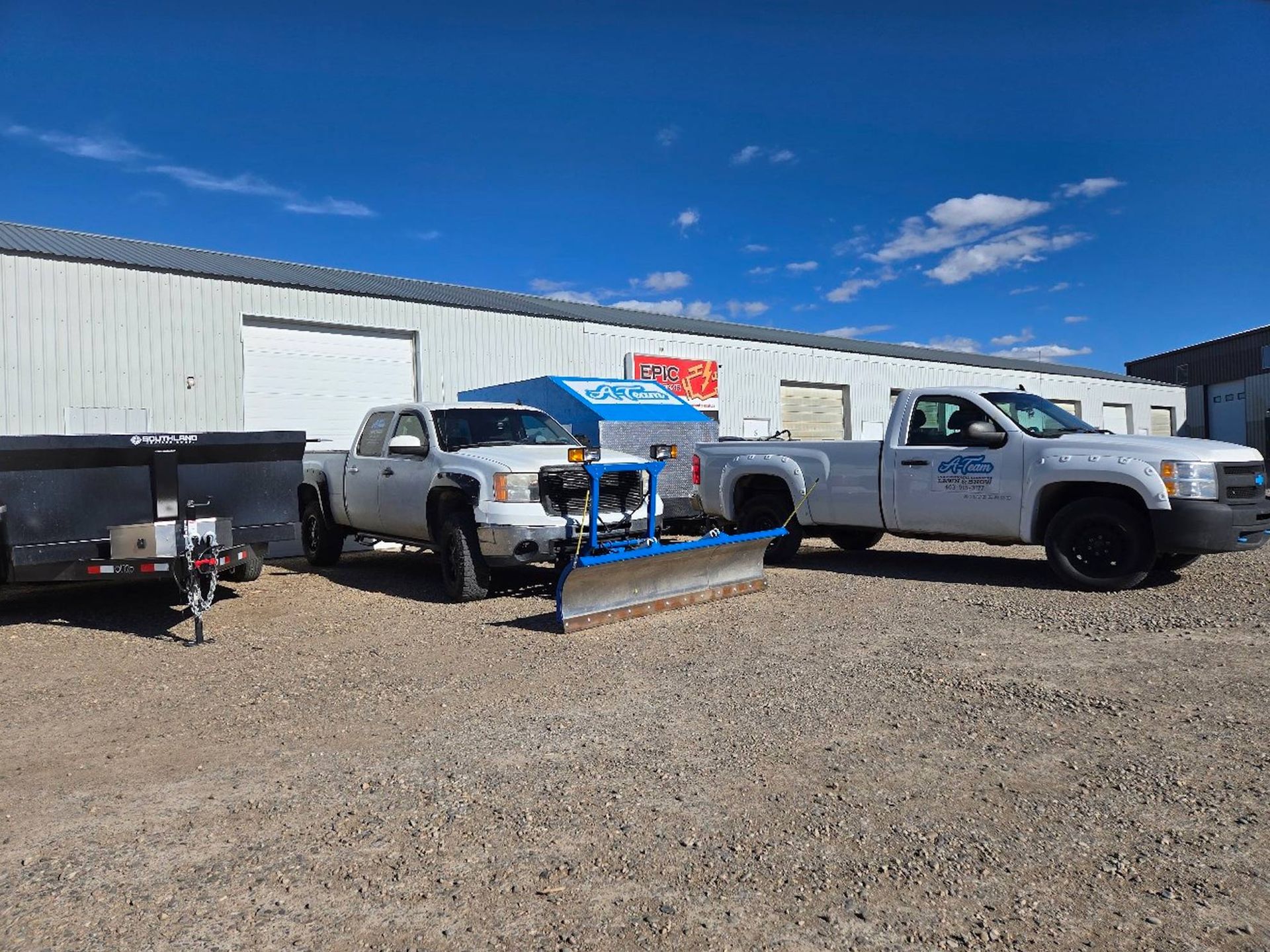 Two white pickup trucks with snowplows parked on a gravel lot in front of a long, white industrial warehouse building.