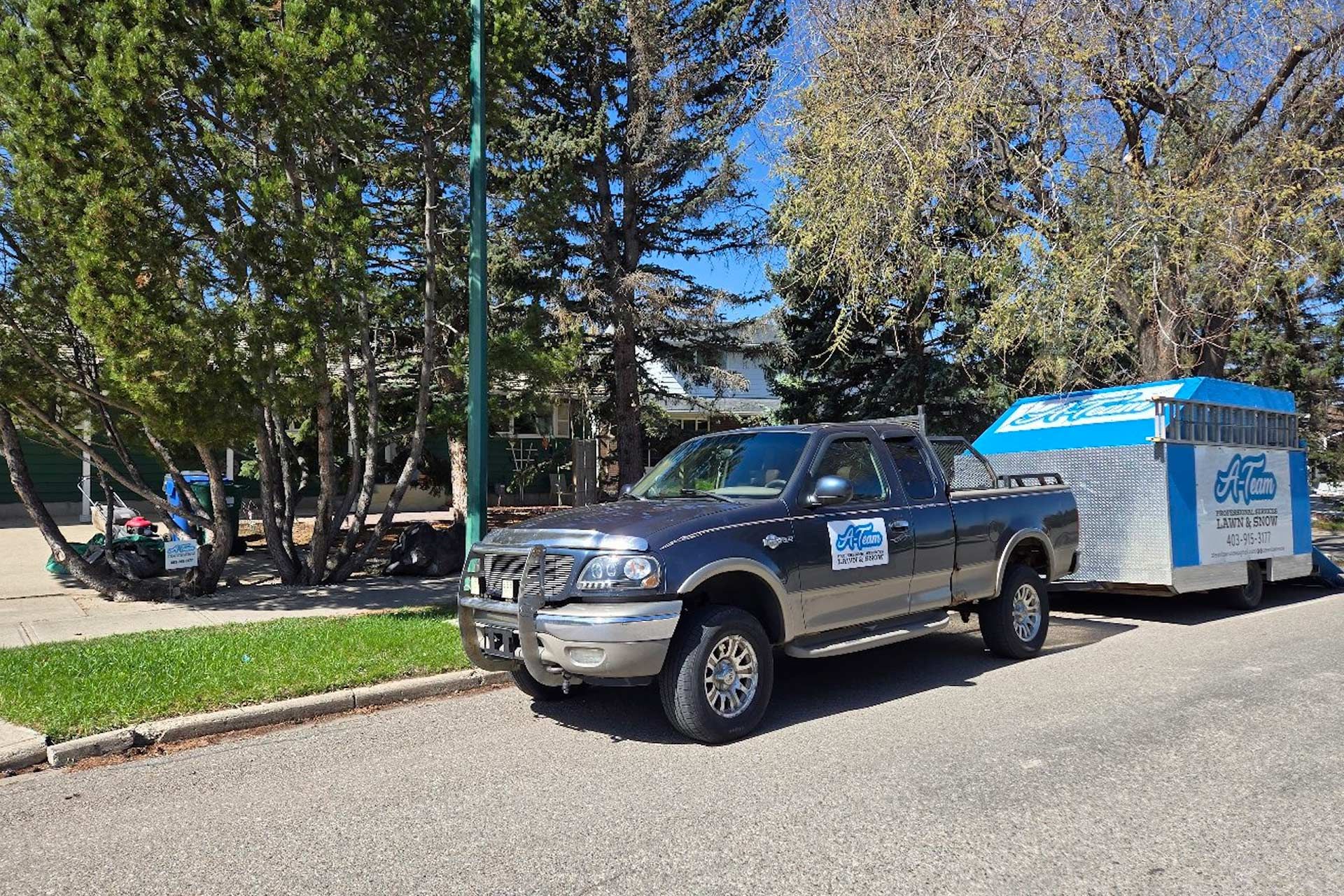 A dark gray pickup truck towing a white and blue cargo trailer is parked on a residential street beside green trees.