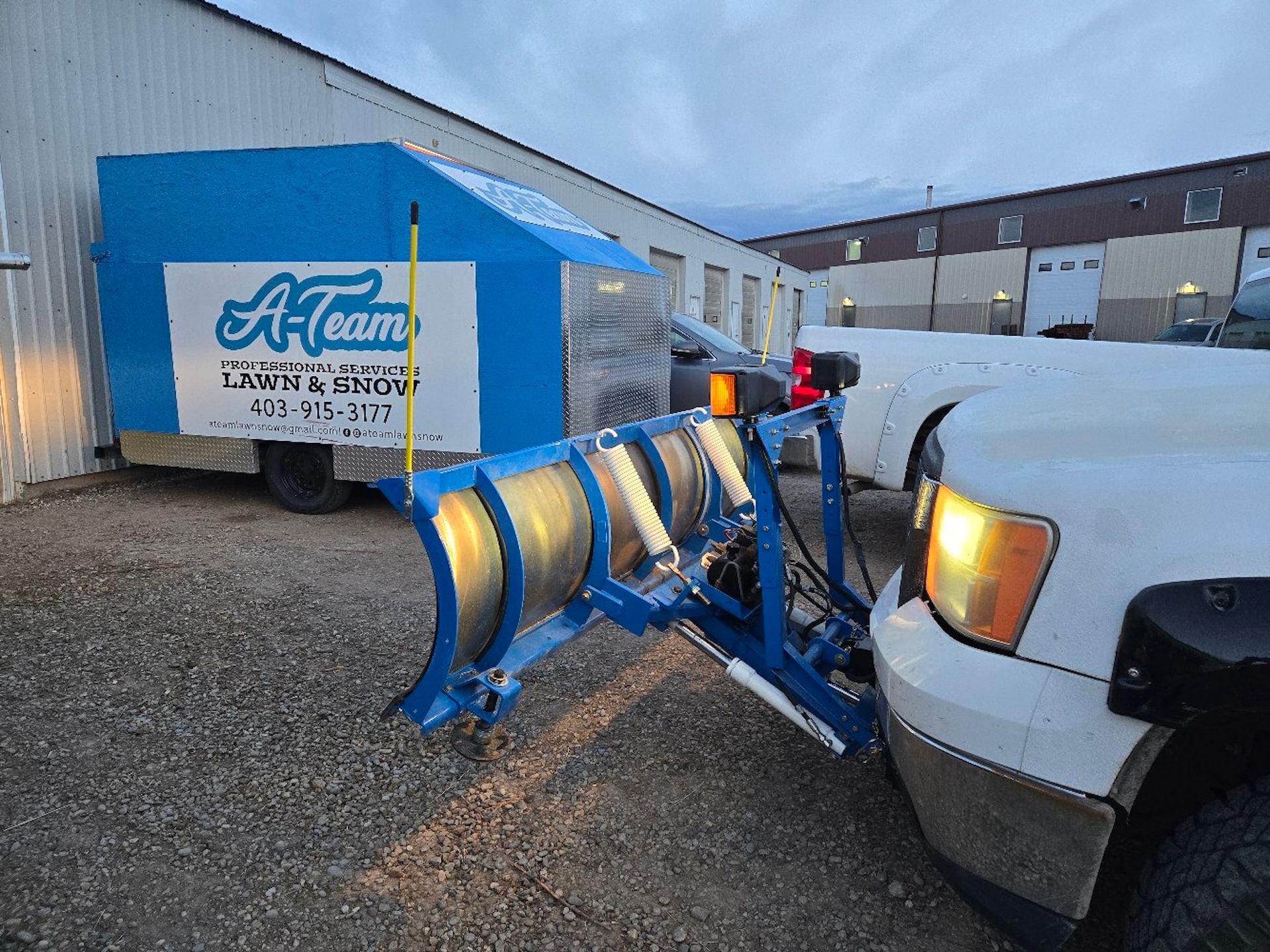 A white pickup truck with a blue snow plow attached, parked next to an 
