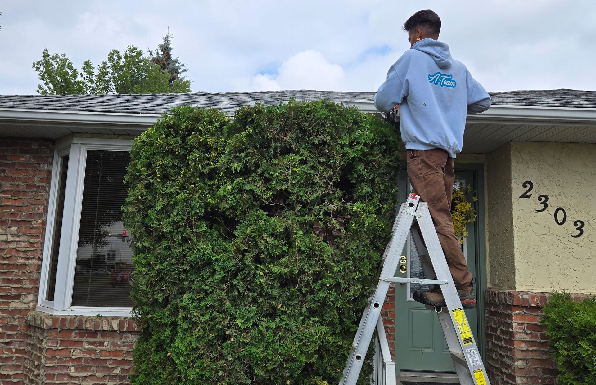 A person in a light blue hoodie stands on a stepladder, cleaning the gutters of a house exterior with brick and stucco.