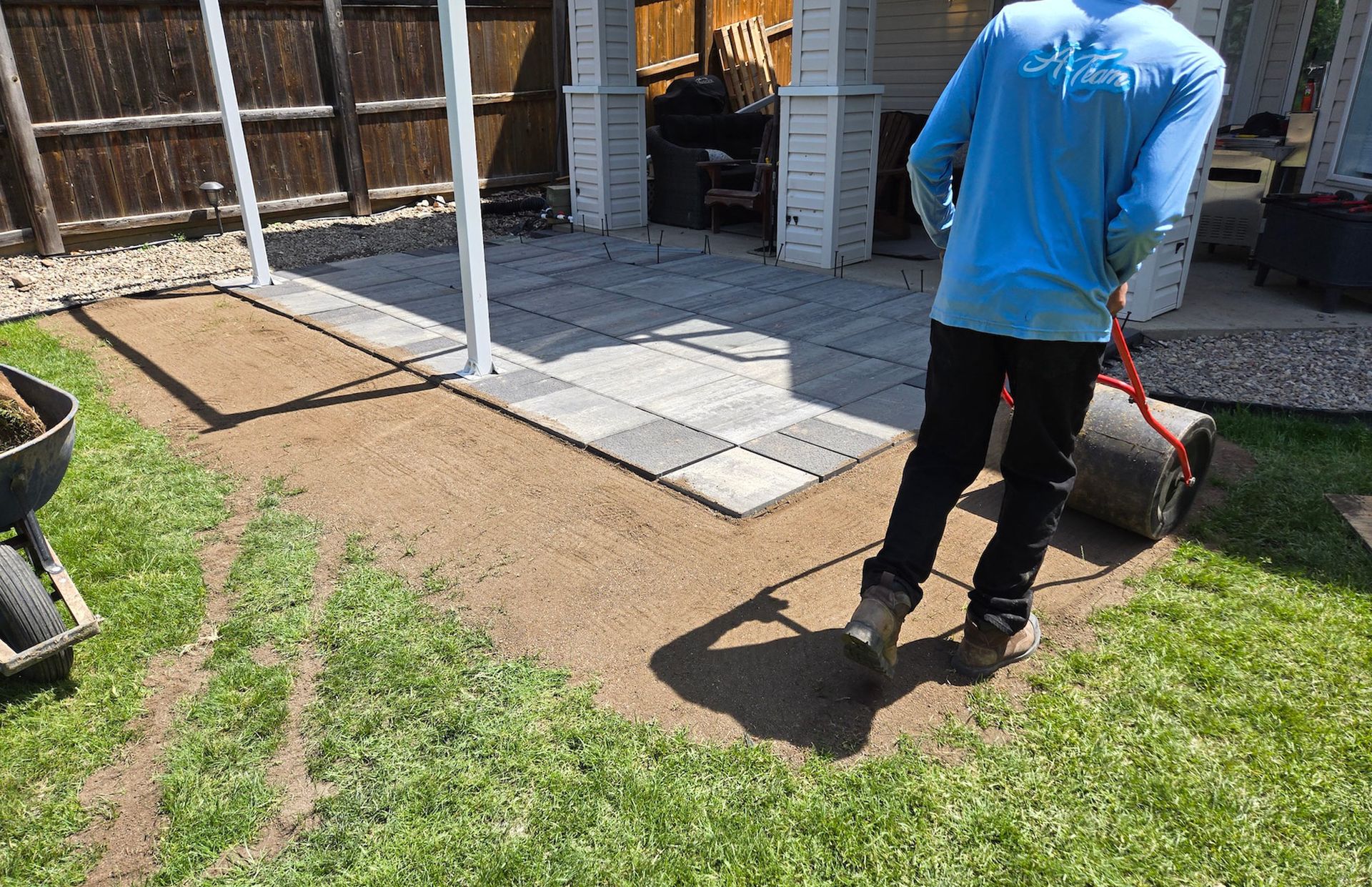 A person in a blue shirt uses a lawn roller to level dirt in a backyard alongside a stone patio.