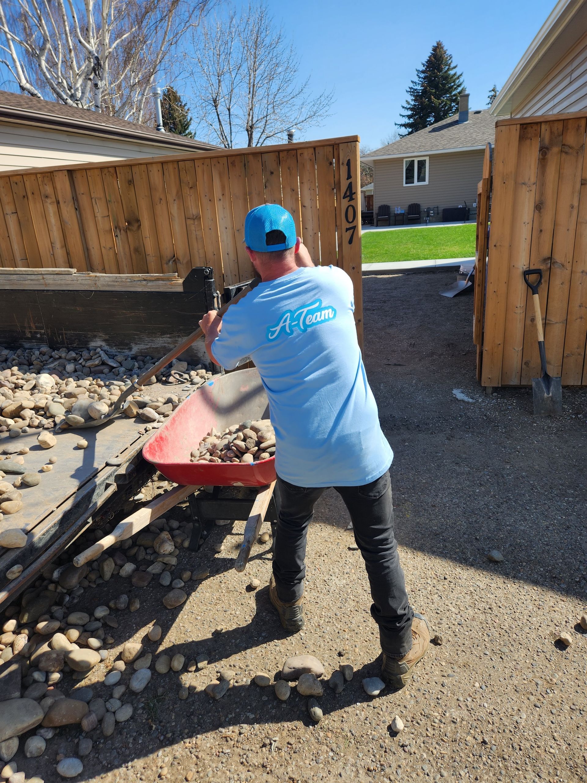 A worker in a light blue t-shirt and blue cap shovels rocks into a red wheelbarrow near a wooden fence.
