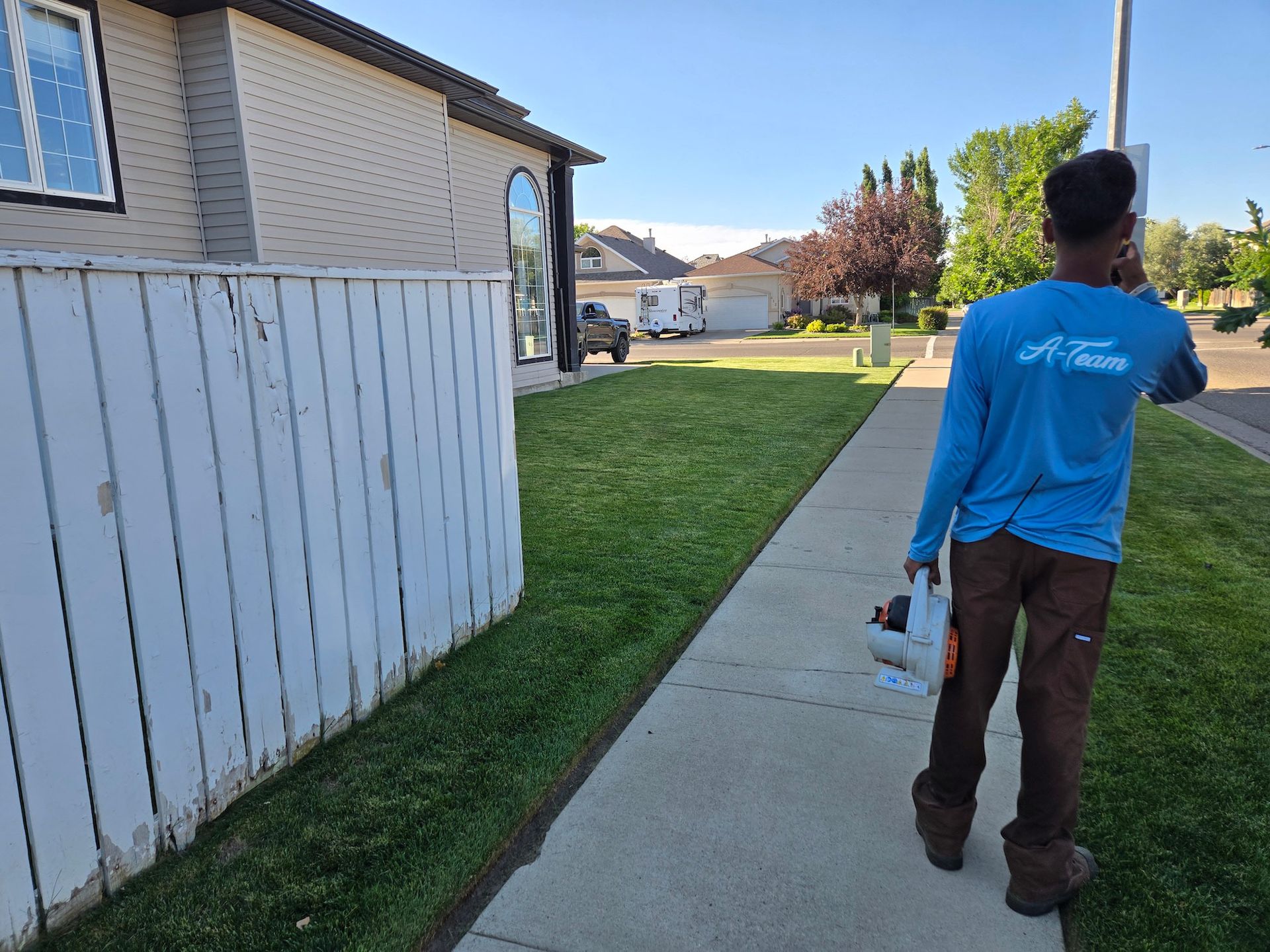 A worker in a blue long-sleeved shirt carries a power leaf blower along a sidewalk next to a house and a white fence.