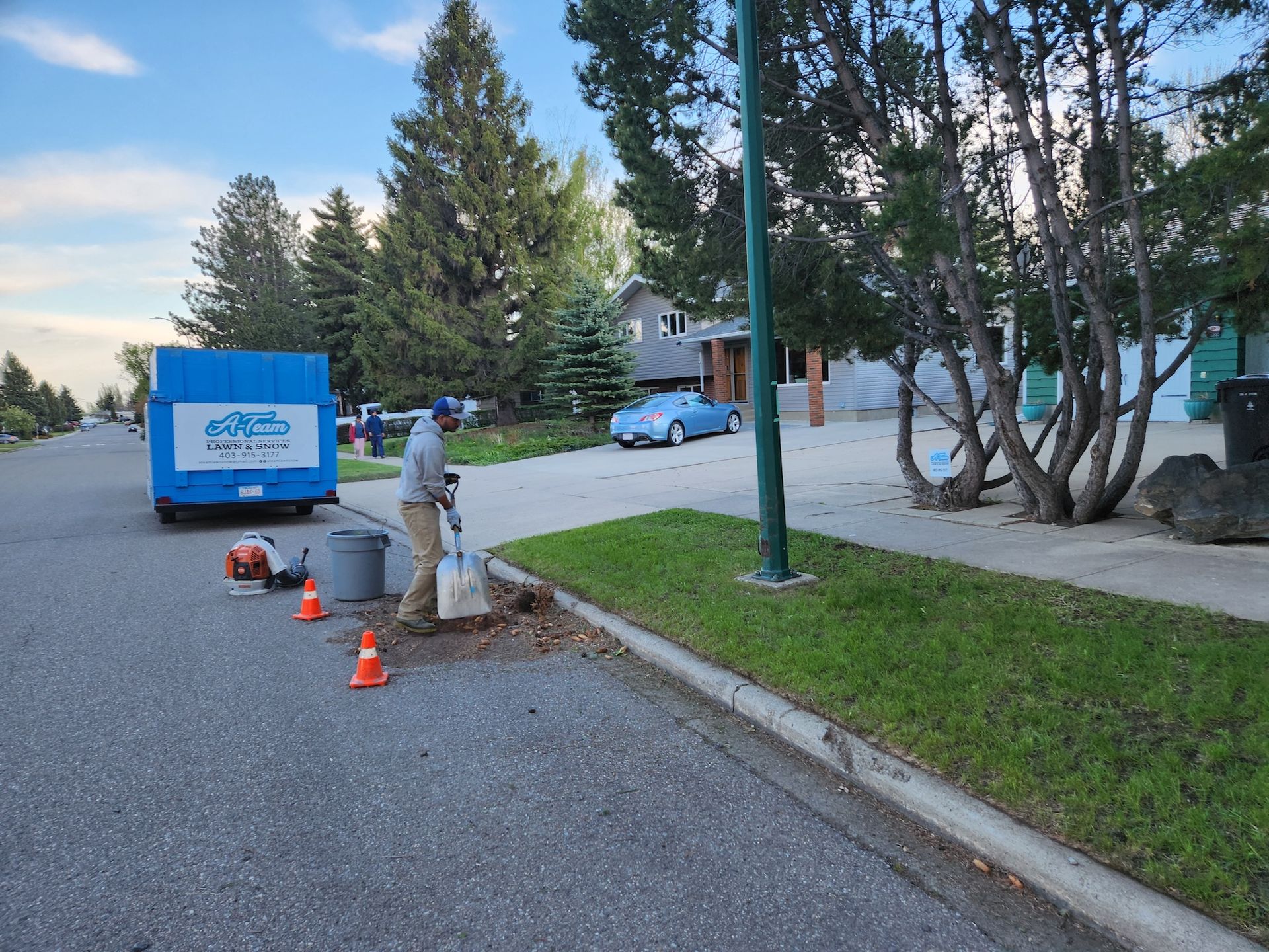 A worker digs at the edge of a residential street near orange traffic cones, a blue dumpster, and a nearby house.