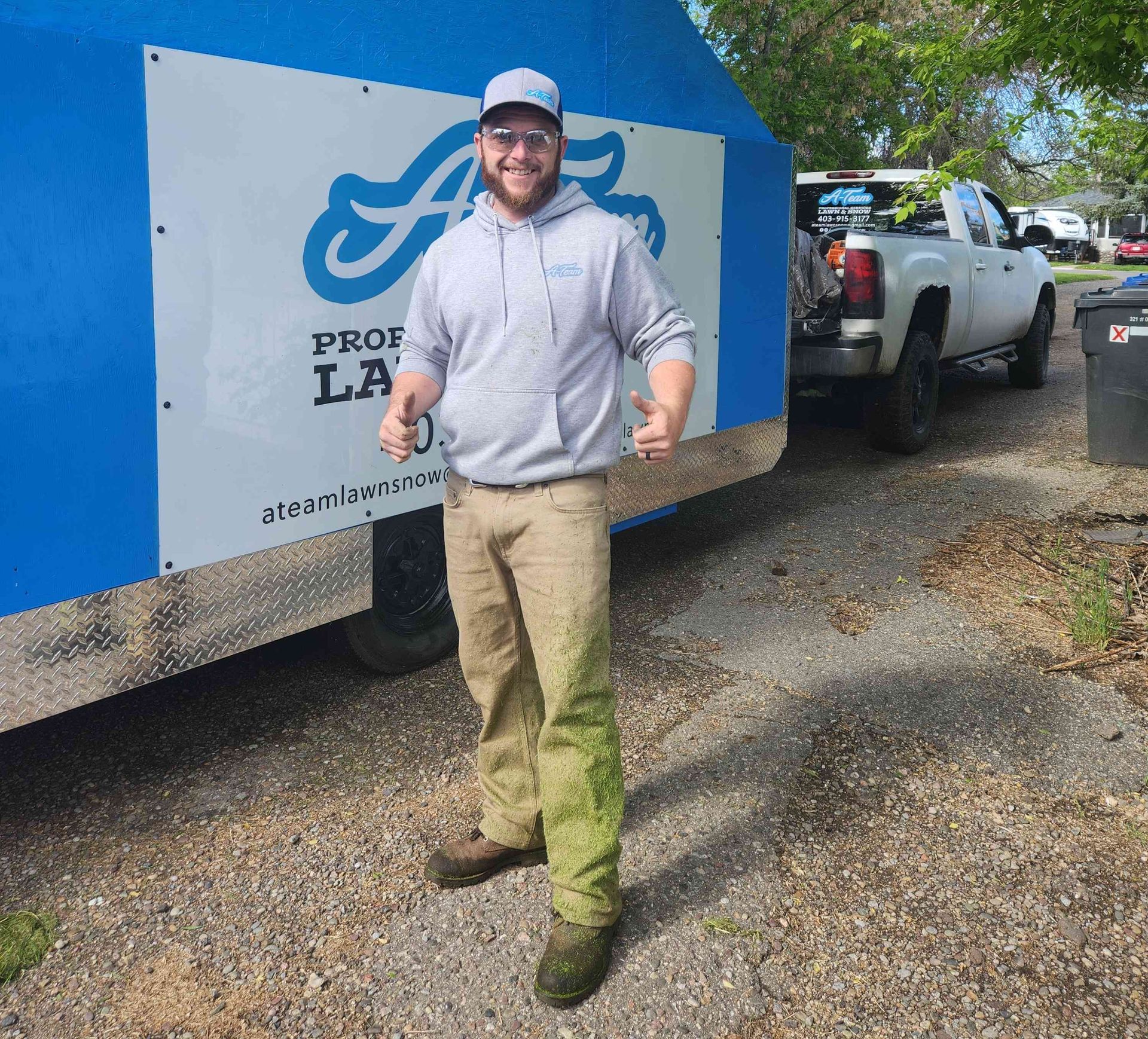A smiling person wearing a grey hoodie, tan pants, and a cap stands giving a thumbs-up in front of a blue lawn care trailer.