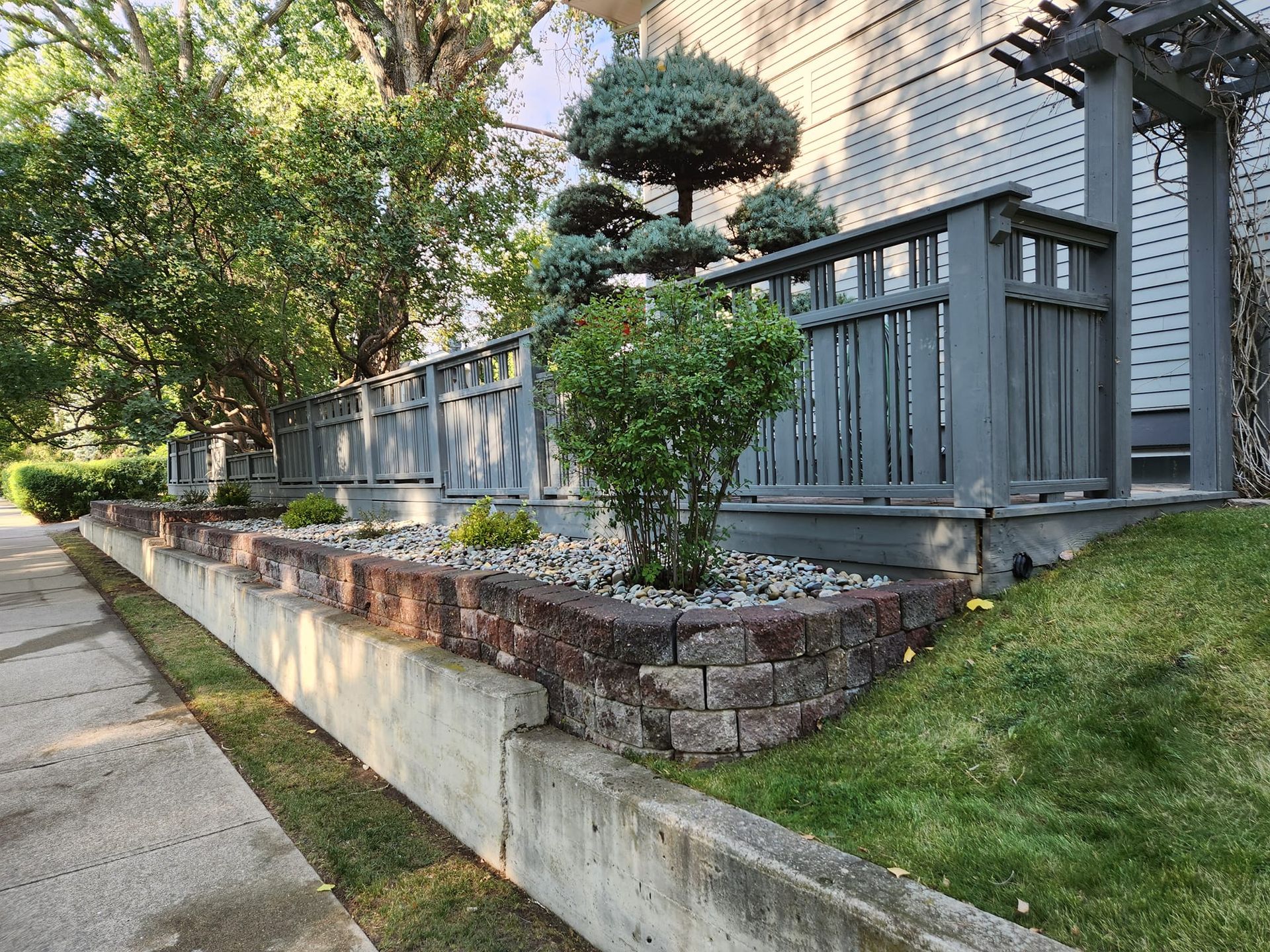 Brick retaining wall with potted plants and a trimmed shrub beside a house porch and sidewalk