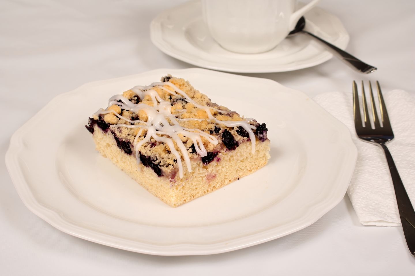 Slice of blueberry crumb cake on a white plate, with a cup and fork.
