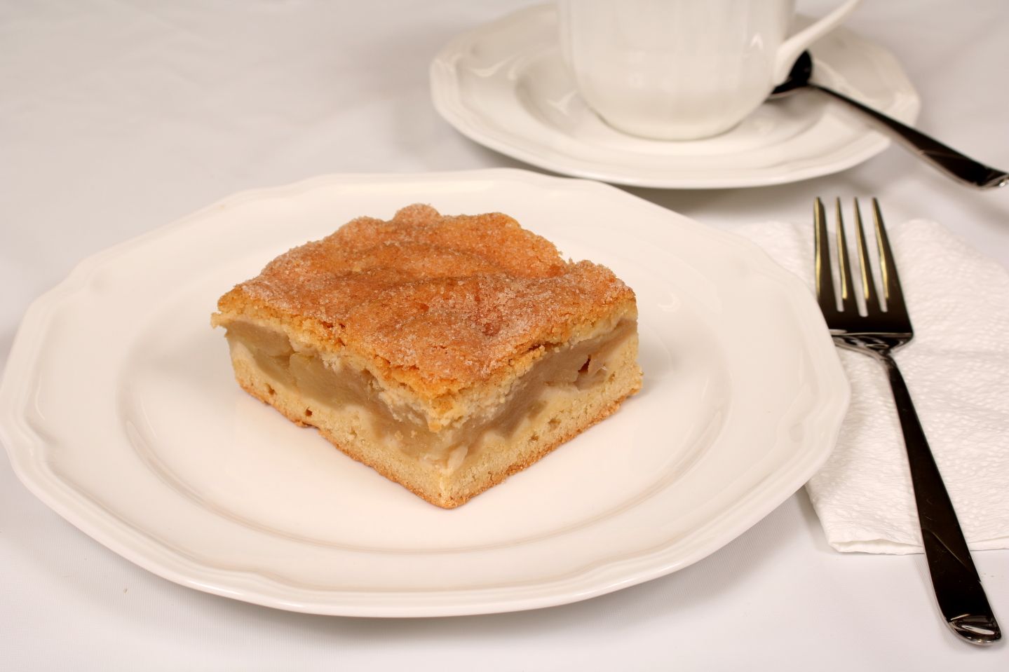Slice of apple cake on a white plate with a fork, napkin, and teacup in the background.