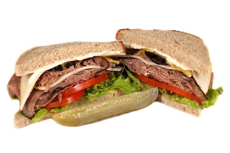 Sliced roast beef sandwich with tomato, lettuce, cheese, and a pickle on a white background.