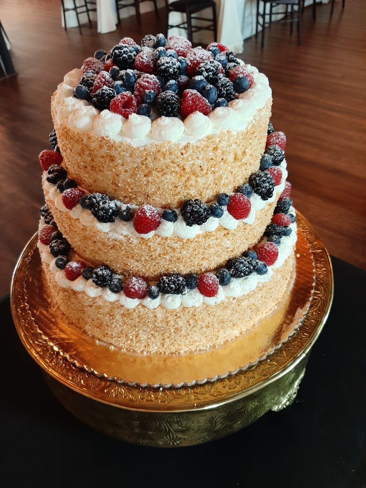 Three-tiered cake with berries on top, on a gold cake stand, set in a wood-floored room.