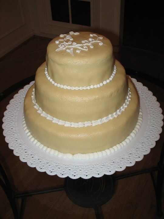 Three-tiered cake with tan frosting, white bead details, and a floral design on top, on a decorative paper doily.