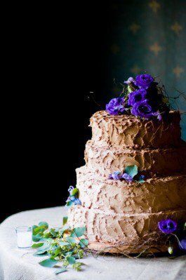 Four-tiered chocolate cake decorated with purple flowers and greenery on a table.