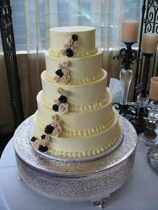 Five-tiered ivory cake on a silver stand, decorated with black and beige flowers, candles in the background.