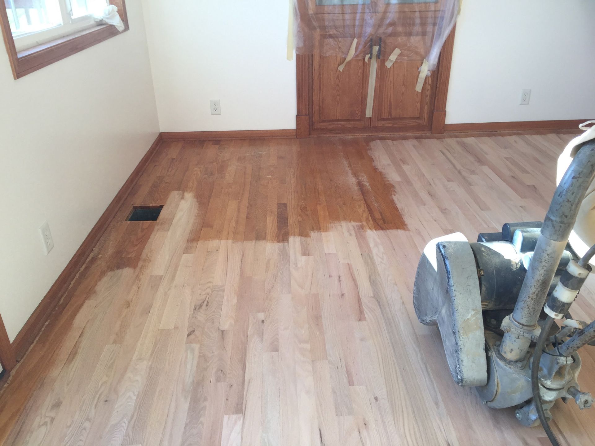 Wood floor being sanded in a small room with a sander and unfinished patches near the door