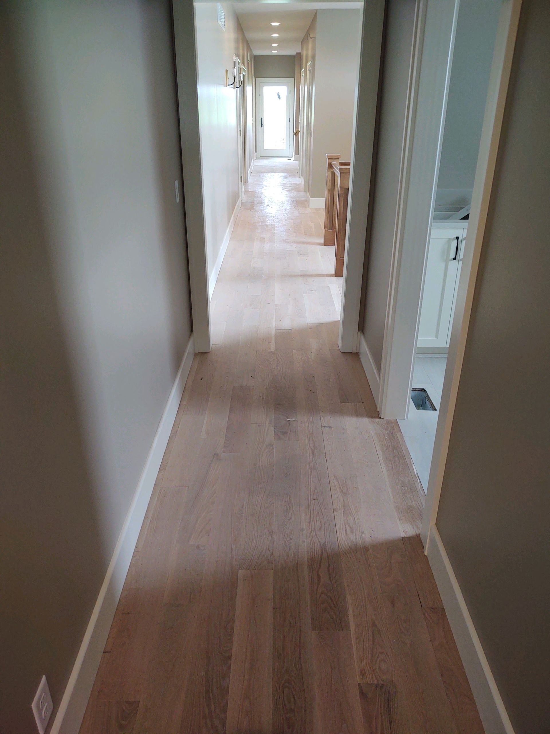Narrow hallway with light wood floors, white walls, and bright windows at the far end
