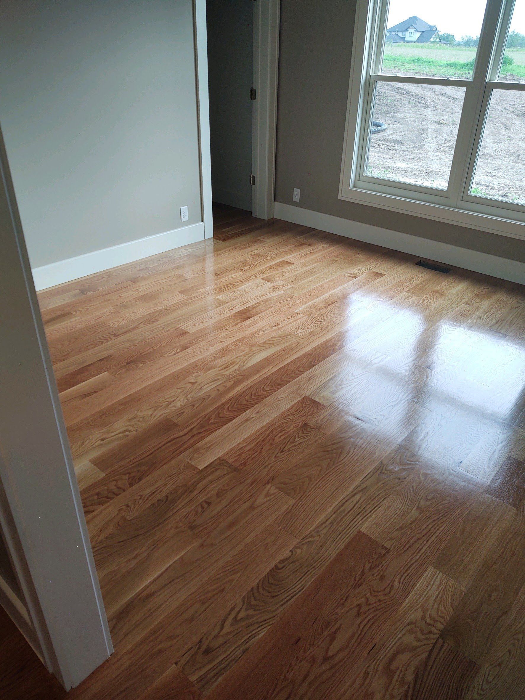 Empty room with glossy hardwood floors and a large window letting in daylight.