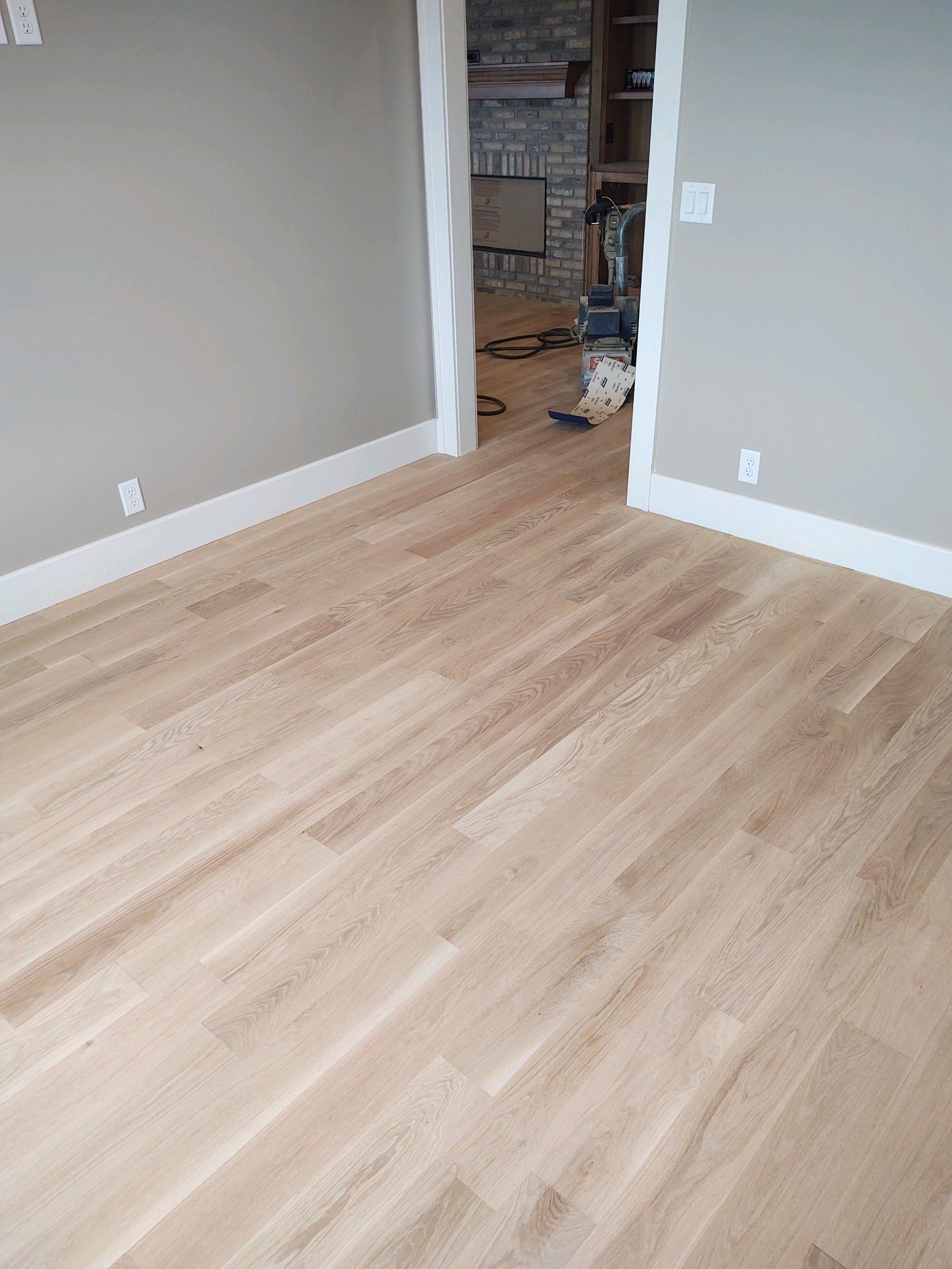 Empty room with light wood floors and gray walls, opening to a cluttered hallway.