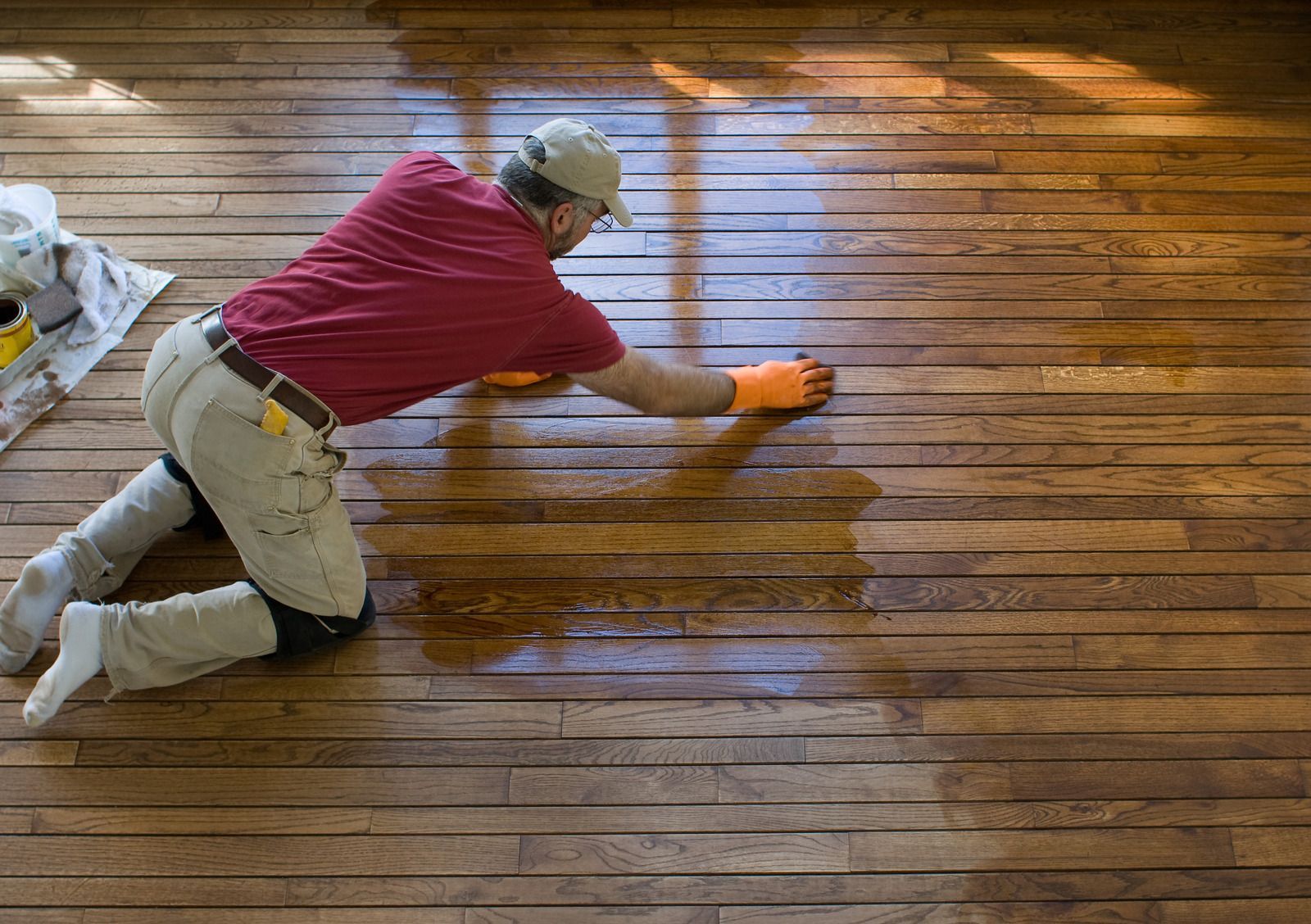 Person kneeling and staining a wooden deck with a brush, wearing a red shirt and beige pants