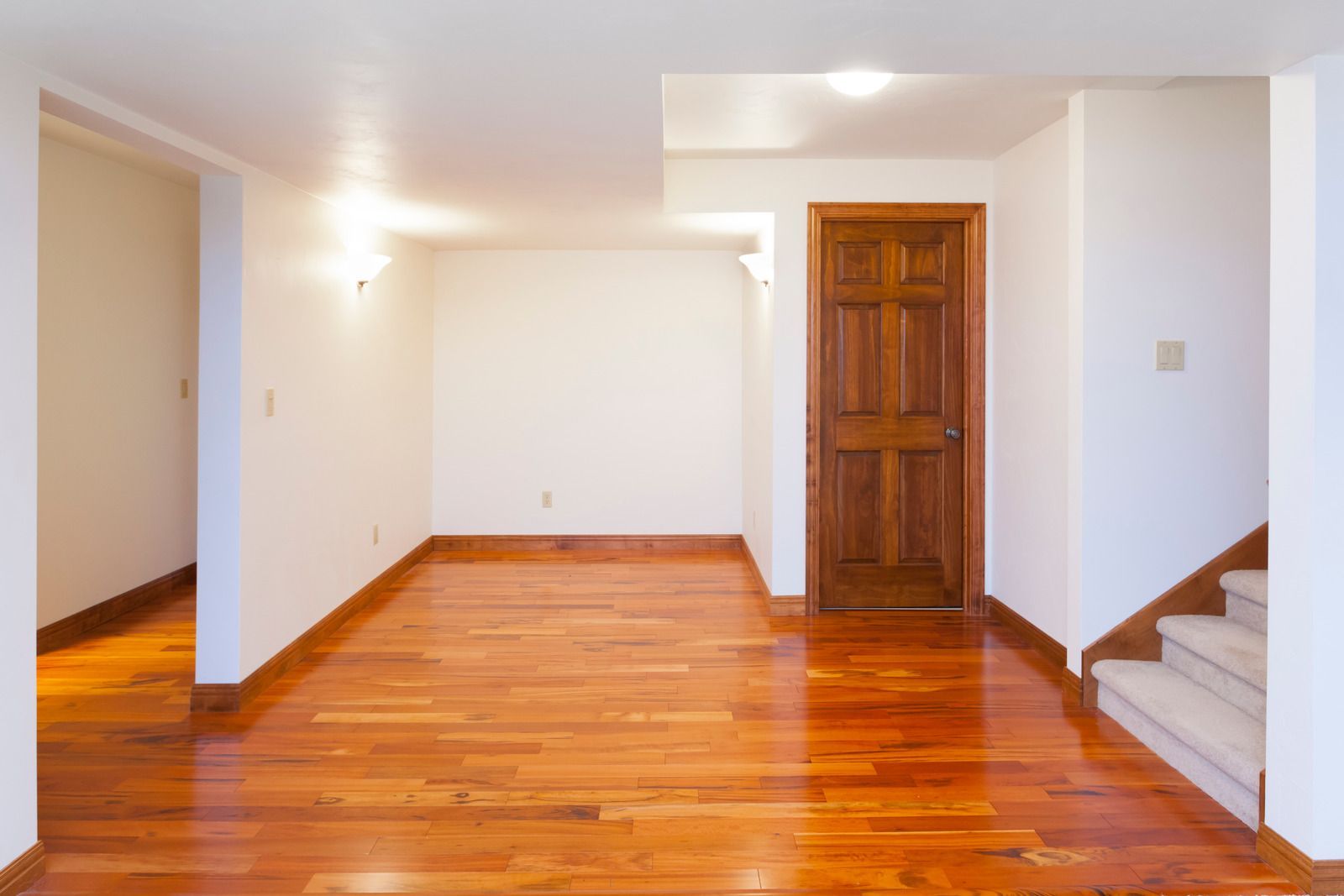 Empty room with glossy hardwood floors, white walls, and a wooden door, viewed from an entryway.