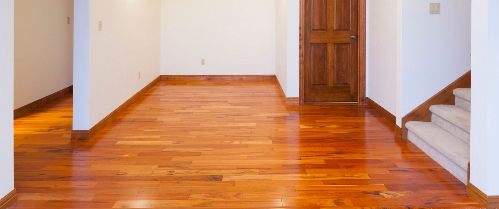 Empty hardwood-floored room with white walls, wooden door, and a staircase on the right.