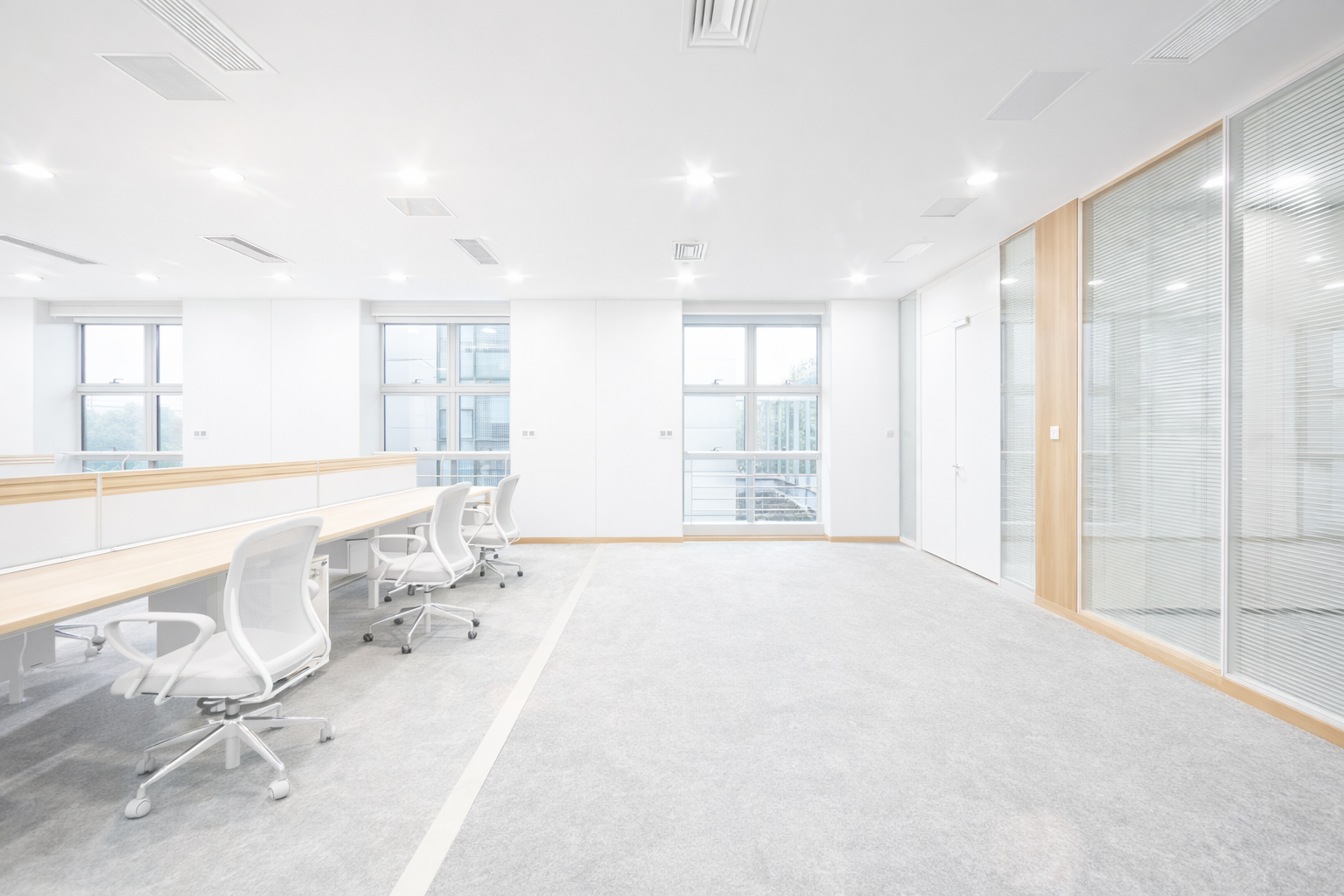 Bright, empty office space with desks, chairs, windows, and light gray carpet.