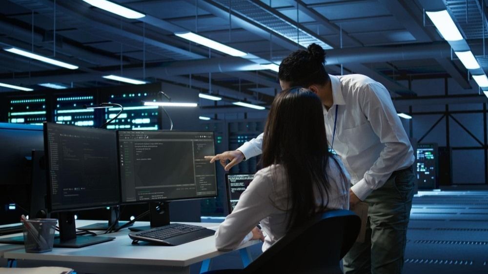 Two people working at computer monitors in a data center, one pointing to the screen.