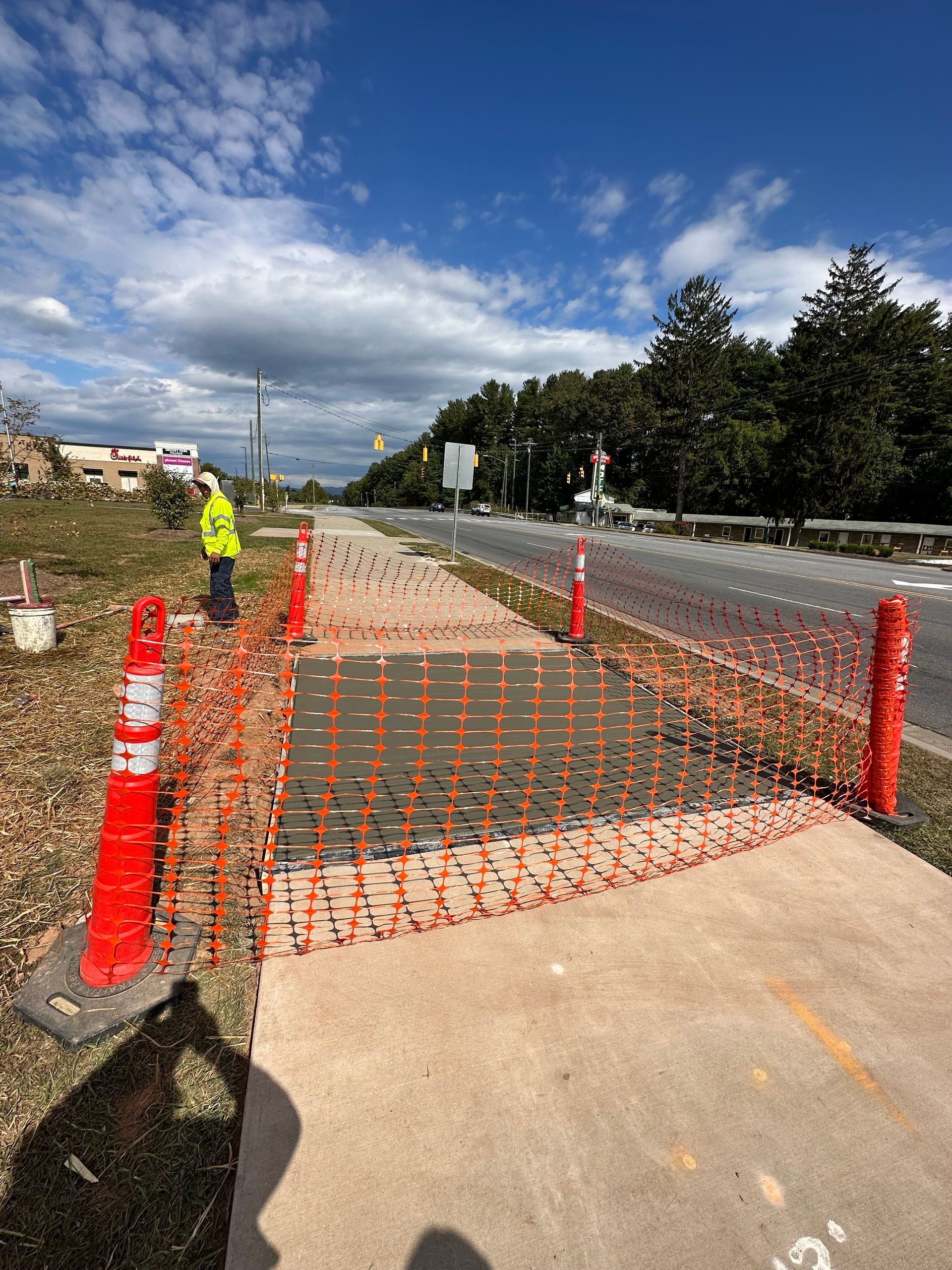A construction worker stands near a sidewalk section cordoned off with orange safety fencing and traffic cones.