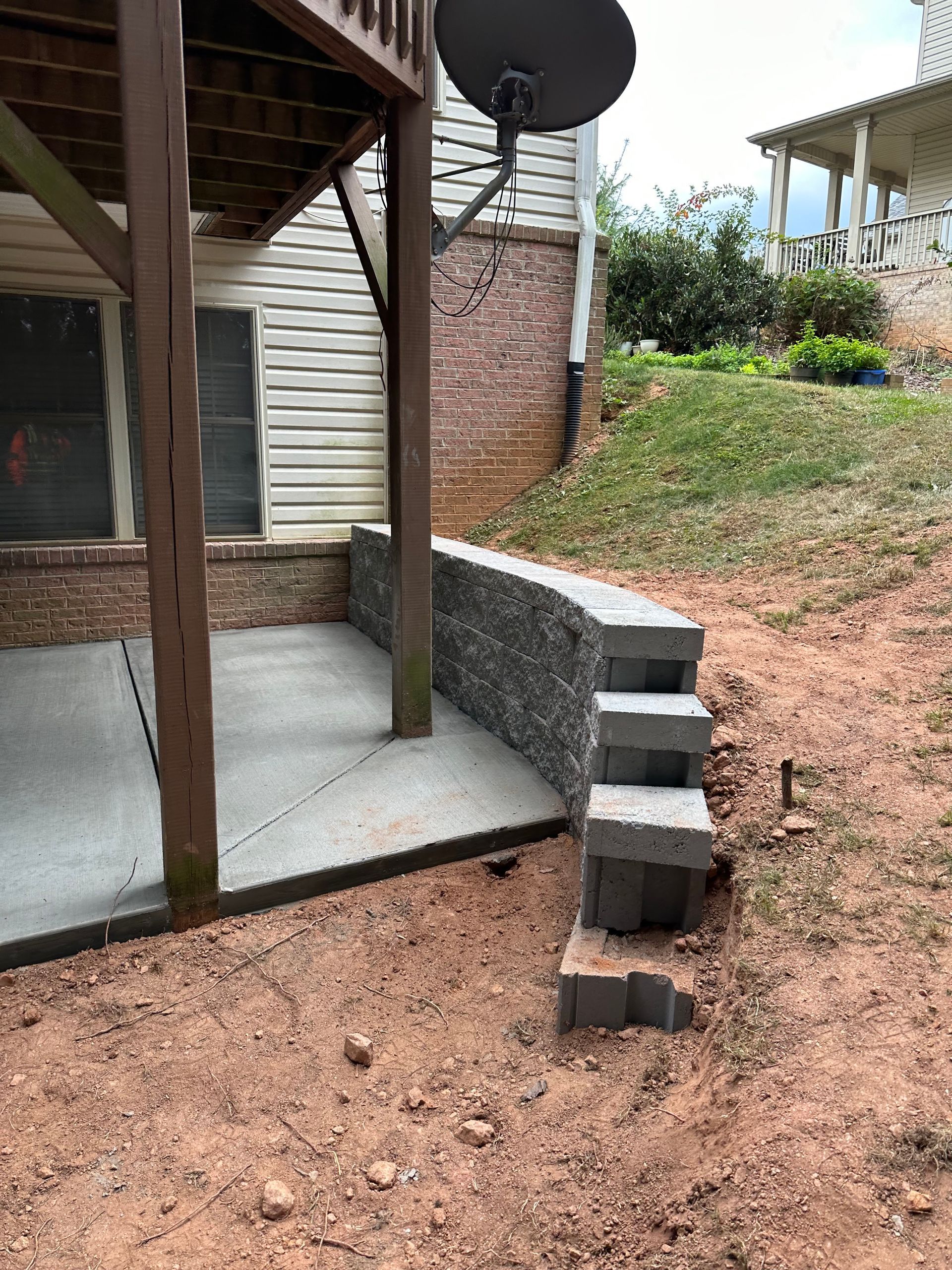 Concrete patio area beneath a wooden deck, featuring a grey stone retaining wall with steps leading up a sloped yard.