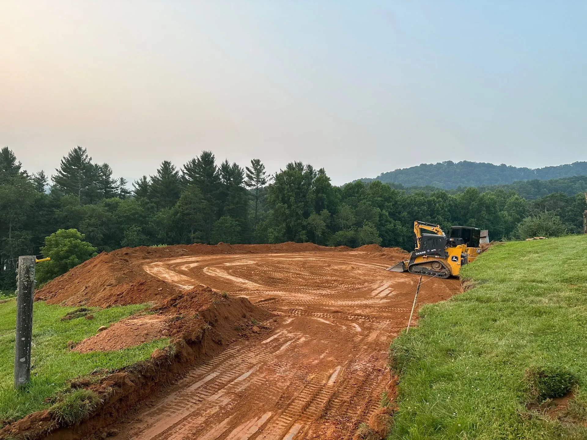 Yellow bulldozer grading a dirt path in a rural field with trees and hills in the background