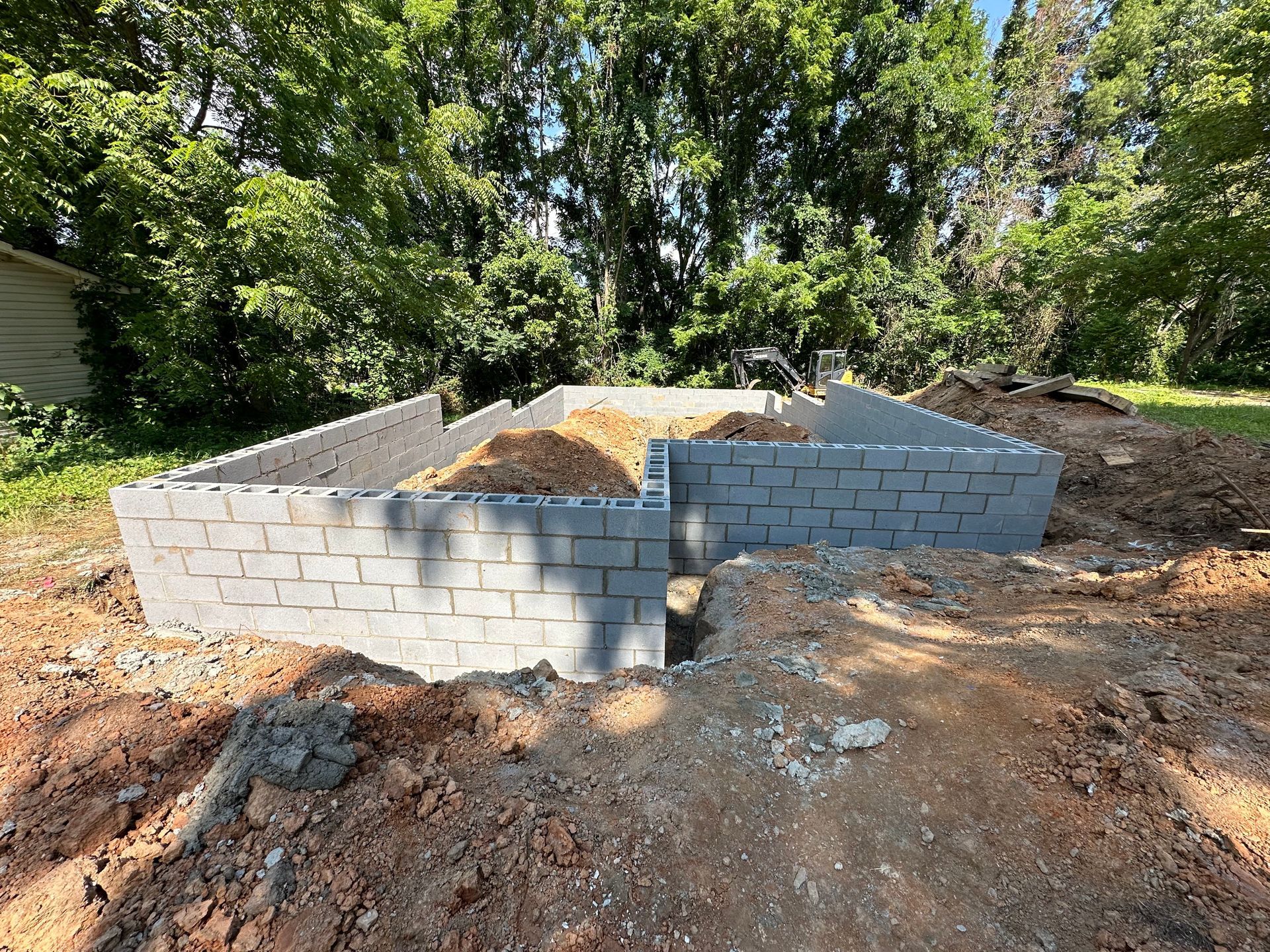 A newly constructed grey concrete block foundation sits in a dirt clearing surrounded by trees on a sunny day.