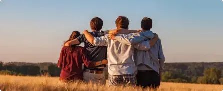 Four friends with arms around each other in a golden field at sunset, facing a distant tree line.