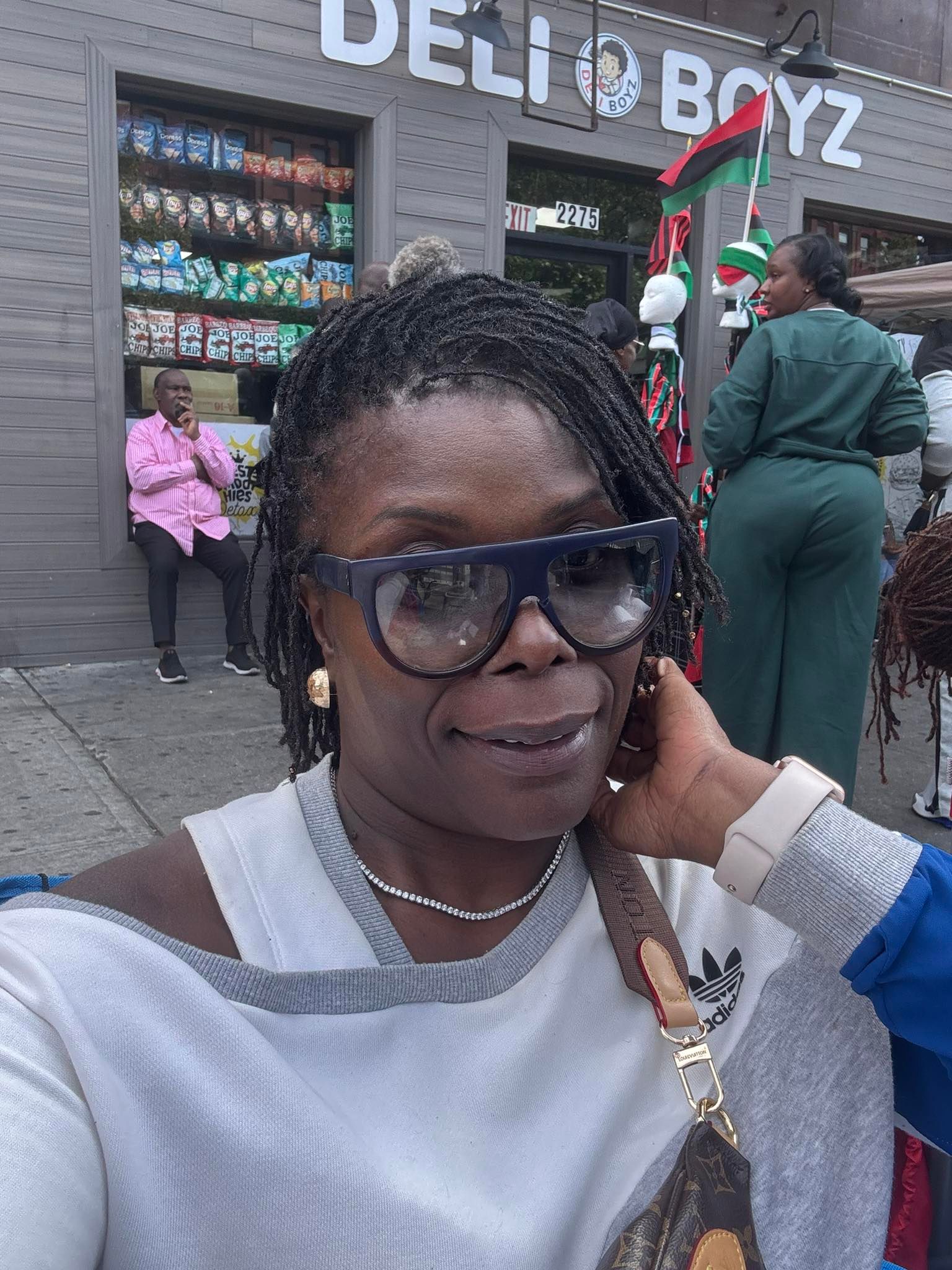 Woman with sunglasses smiles in front of a deli. Background features another person, building facade.