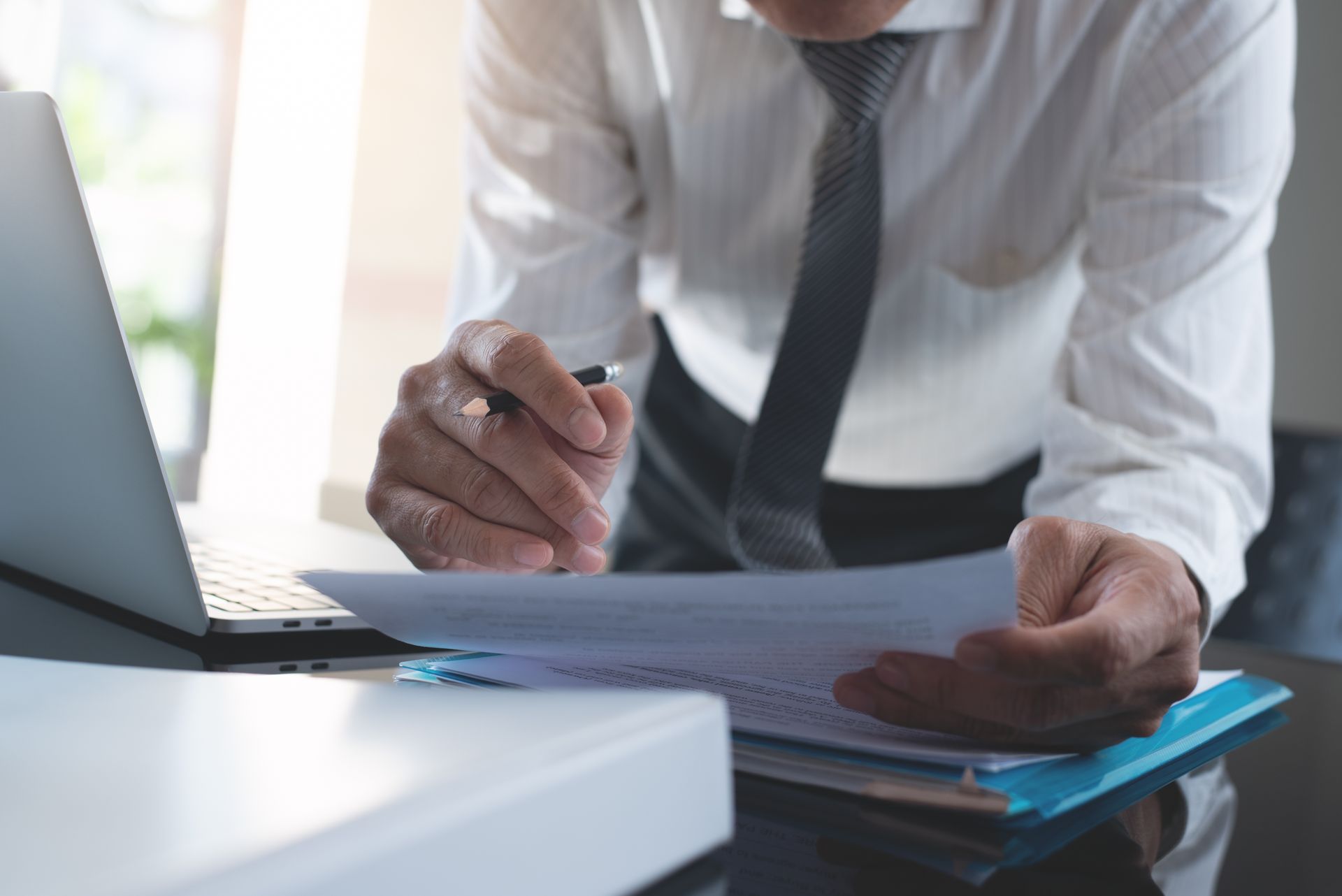 Man in white shirt and tie examines documents, leaning over a laptop on a desk.