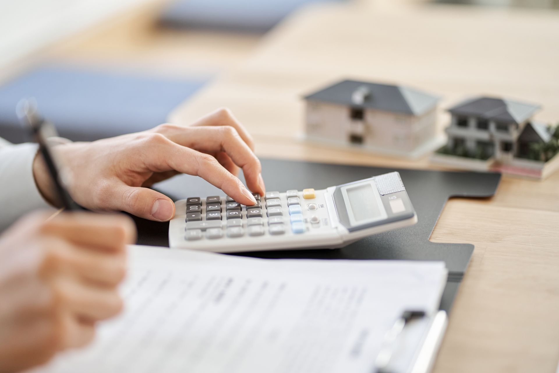Hands using calculator over paperwork, two model houses in the background.