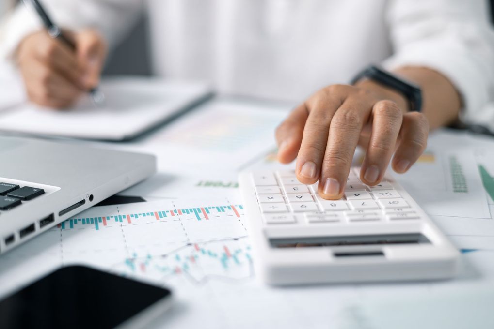 Person using a calculator and taking notes, with a laptop, smartphone, and financial charts on a desk.