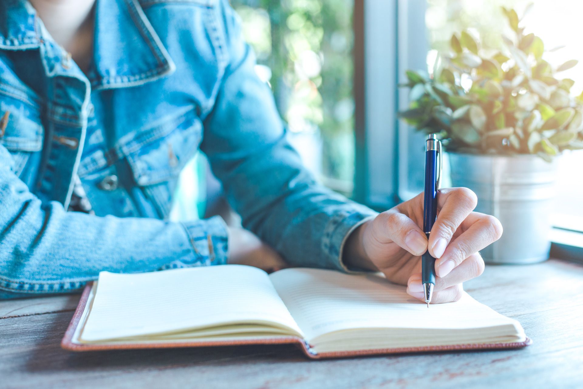 Person writing in an open notebook at a table by a window, wearing a denim jacket.