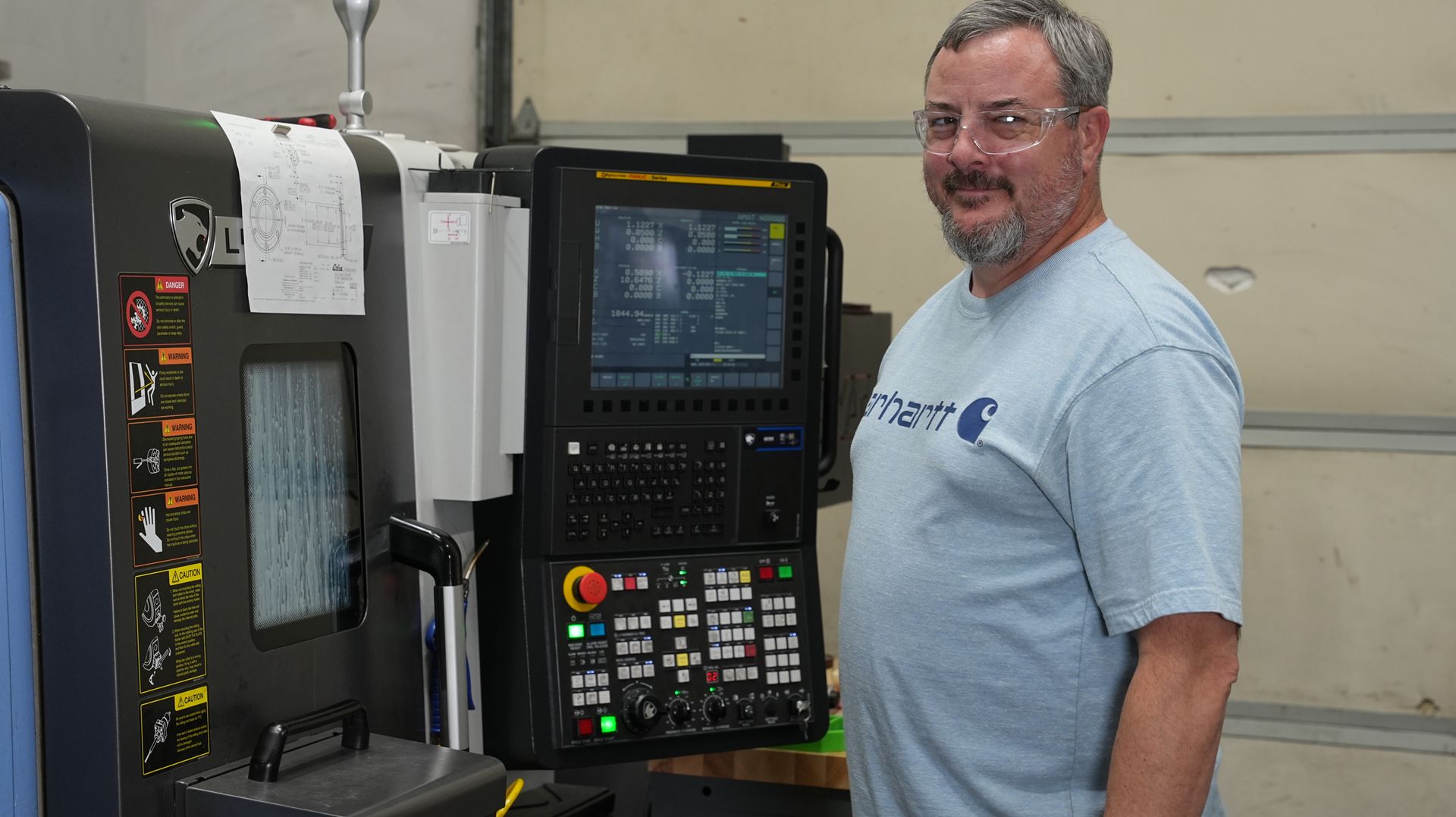 A man with a beard is smiling while looking at the control screen of a CNC machine. 