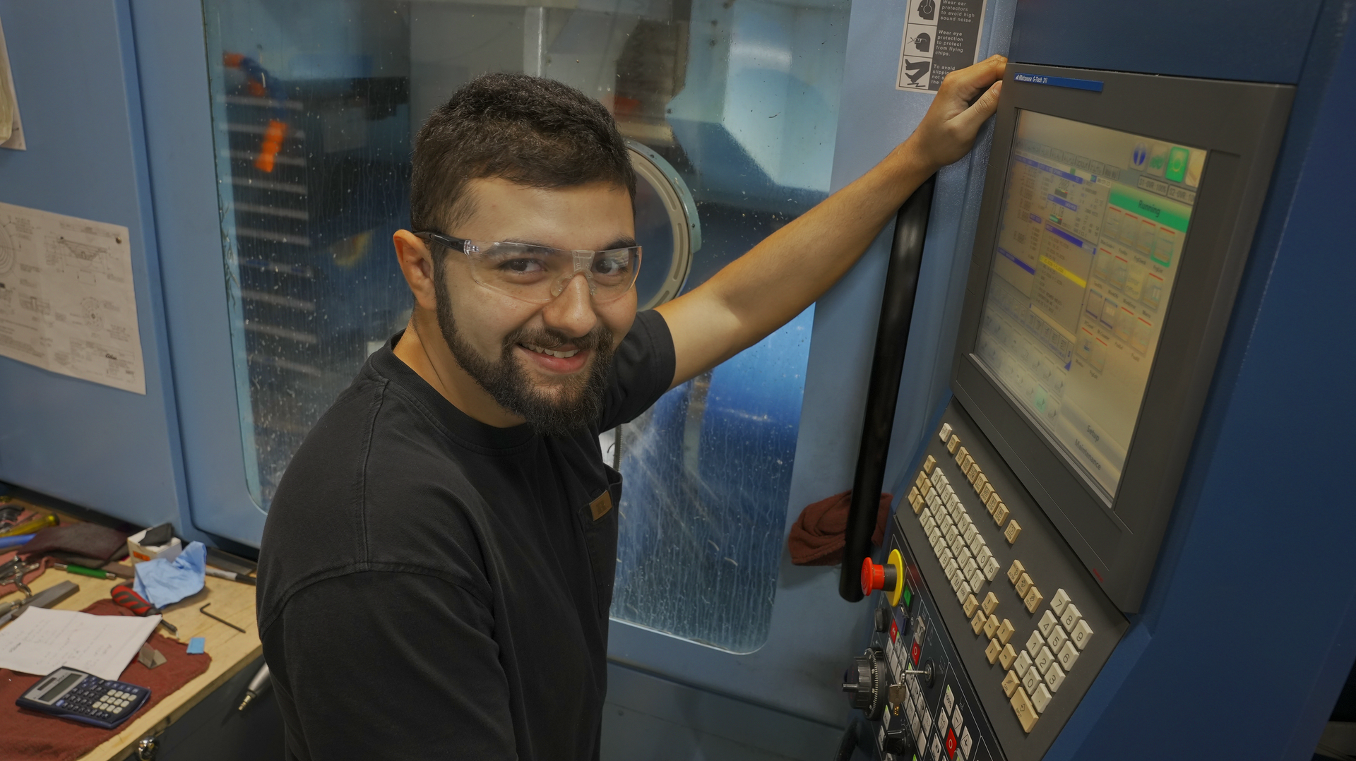 A man with a beard wearing protective eyeware is standing in front of a 5-axis Matsura machine.