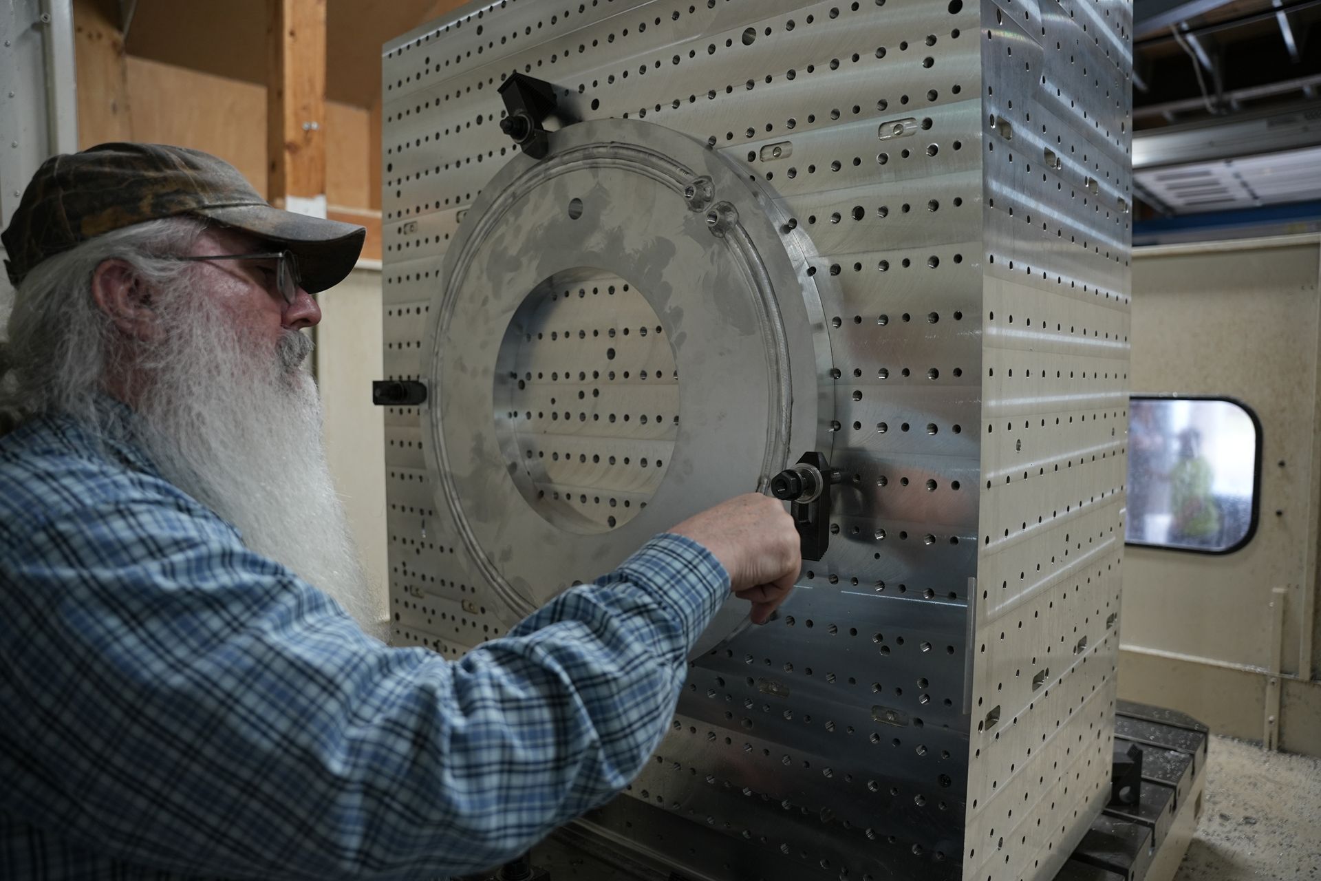 A man with a long beard and baseball cap is loading a product onto the Shibaura cutting machine.