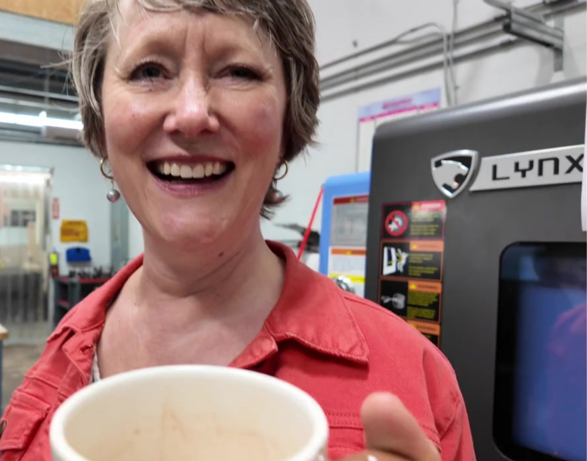 A smiling woman in an orange jacket stands in front of a CNC machine in a factory.