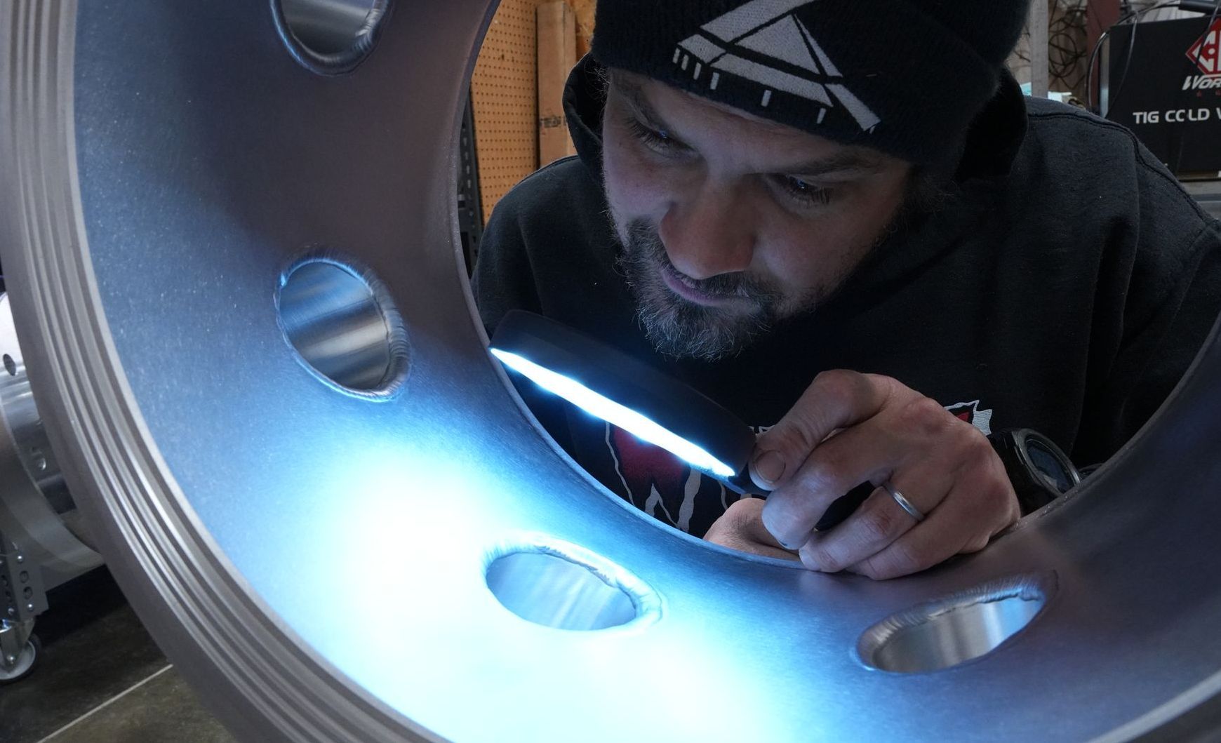 A man is inspecting the inner wall of a large drum shaped aluminum vacuum chamber using a magnifying glass. 