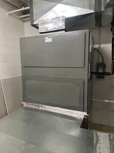 Gray HVAC unit against a white brick wall, with ductwork above and metallic floor in a basement.
