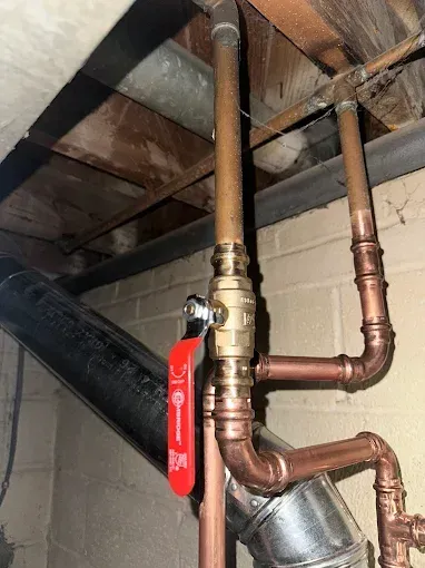 Copper pipes and a brass ball valve with a red handle are installed in a basement, with a black pipe visible.