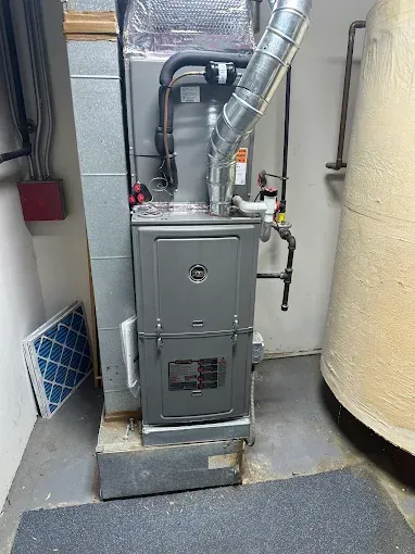 A furnace in a basement setting. Grey metal exterior, connected to ductwork, pipes and water heater. Filter visible.