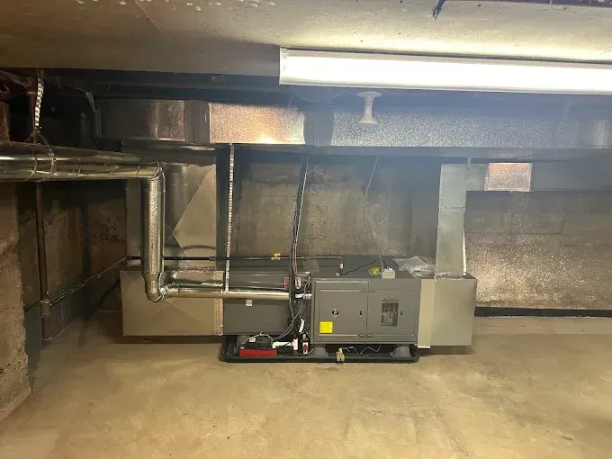 Furnace in basement with ductwork. Gray metal unit on concrete floor. Fluorescent light above.