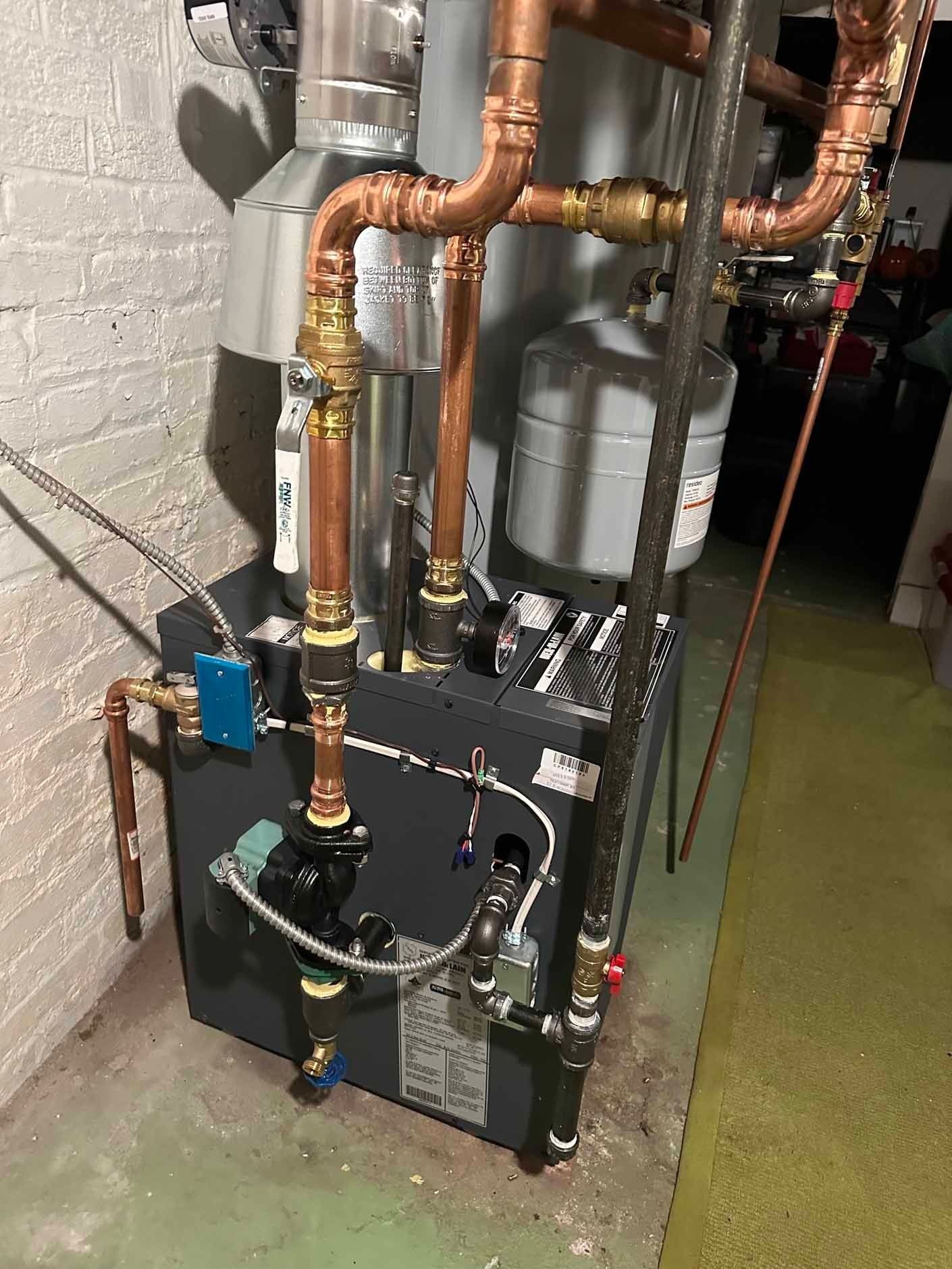 A dark gray boiler with copper pipes and a silver expansion tank in a basement setting.